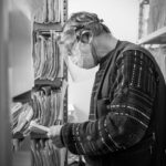 Masked archivist sorts paper files on crowded storage shelves