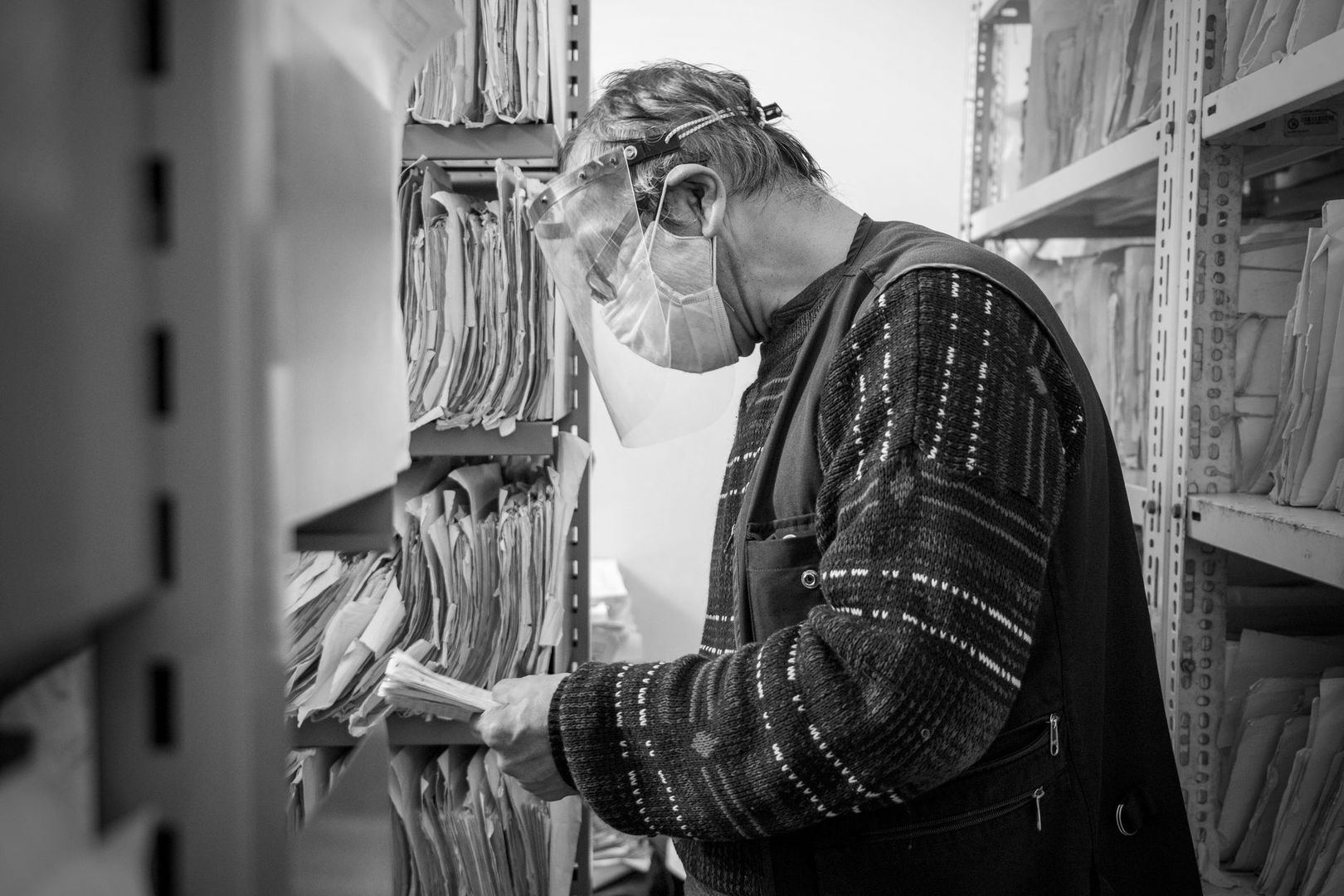 Masked archivist sorts paper files on crowded storage shelves